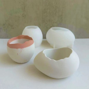 Pode incluir: Quatro vasos de cerâmica branca de diferentes formas e tamanhos. Um vaso tem uma borda rosa claro.