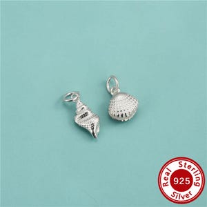 Könnte beinhalten: Zwei silberne Anhänger in Form von Muscheln, jeweils mit einem kleinen runden Ring an der Oberseite zur Befestigung. Ein Anhänger ist eine spiralförmige Turmschnecke, der andere eine abgerundete Muschel, beide mit detaillierten Texturen.
