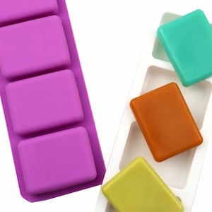Pode incluir: Dois moldes de silicone para fazer barras de sabão. O molde à esquerda é roxo e tem quatro cavidades retangulares. O molde à direita é branco e tem quatro cavidades retangulares. Existem quatro barras de sabão no molde branco, uma de cada cor: verde, laranja, amarelo e branco.