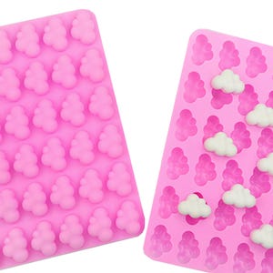 May include: Two pink silicone moulds. One is filled with small, three-lobed shapes. The other has the same shapes, plus white cloud-shaped sweets. These moulds are likely for making sweets, chocolates, or other treats.
