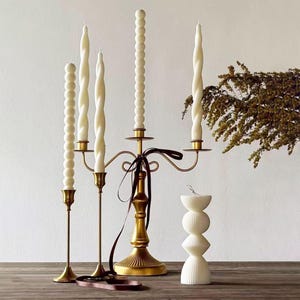 Peut inclure: Une collection de bougies décoratives et de chandeliers. L'image présente un candélabre doré avec cinq bougies blanches en spirale et perlées. Deux autres bougeoirs dorés avec des bougies blanches sont également présents, ainsi qu'une bougie empilée unique.