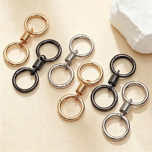May include: A collection of six metal keychains, each featuring two circular rings connected by a central cylindrical spring mechanism. The set includes gold-toned, silver-toned, and black-toned finishes, all with a smooth, polished metallic surface.