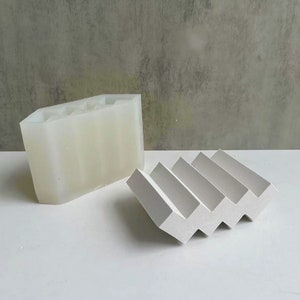 May include: A white soap dish with a zig-zag design and a translucent, rectangular mold with vertical slots. The soap dish is made of a solid material, and the mold is made of a flexible material. The background is a neutral grey.