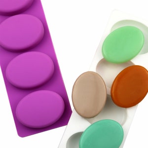 Puede incluir: Dos moldes de silicona para hacer barras de jabón ovaladas. Un molde es morado y tiene cuatro cavidades. El otro molde es blanco y tiene seis cavidades. Hay tres barras de jabón en el molde blanco: una es verde claro, una es naranja y una es marrón claro.