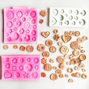 Op de afbeelding: Twee roze siliconen mallen voor het maken van knoopvormige decoraties. De mallen hebben verschillende vormen, waaronder harten, sterren, cirkels en vierkanten. Er zijn ook veel goudkleurige knopen verspreid rond de mallen.