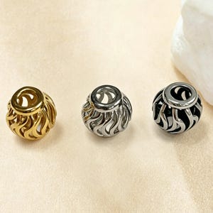 Puede incluir: Tres cuentas esféricas con un patrón de llamas recortado. Una cuenta es de tono dorado, otra de tono plateado y otra de tono gris oscuro metalizado. Cada cuenta presenta una abertura circular lisa en la parte superior y un diseño de celosía hueco.