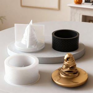 Peut inclure: Un ensemble de moules pour la fabrication artisanale, dont un Choixpeau magique doré, un récipient cylindrique noir à motif de briques et deux autres moules cylindriques blancs.