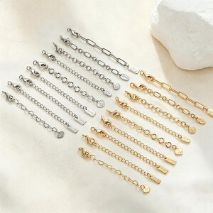 May include: A collection of silver-toned and gold-toned metal jewellery extender chains, each featuring a lobster claw clasp at one end and a small decorative charm or tag at the other. The chains vary in link style, including oval, round, and heart-shaped designs.
