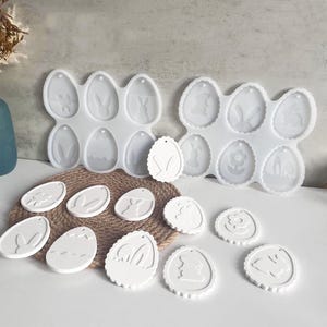 May include: White silicone moulds for Easter crafts. The moulds feature egg shapes with bunny and floral designs. Several finished white resin Easter egg ornaments are also displayed, showcasing the potential of the moulds for DIY projects.