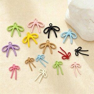 Puede incluir: Una colección de pequeños dijes con forma de lazo en varios colores, incluyendo rosa, verde, amarillo, negro, azul, morado, beige, rojo y blanco. Cada dije cuenta con una anilla en la parte superior para colgar, con un acabado mate y liso.