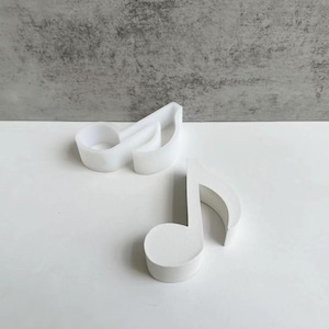 Puede incluir: Dos formas blancas de notas musicales, una un molde y la otra un producto terminado, se encuentran sobre una superficie blanca. El molde tiene forma de clave de sol. El producto terminado es una negra. El fondo es gris texturizado.