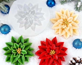 Puede incluir: Un molde de silicona transparente con forma de flor de pascua junto a tres flores de pascua de colores crema, verde y rojo. Las flores tienen centros dorados y están sobre una superficie nevada con adornos azules y agujas de pino.
