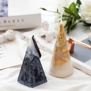 May include: Two decorative candles in the shape of a pyramid and a cone. The pyramid candle is blue and white with a label that says "MAN YUE". The cone candle is white with gold leaf accents.