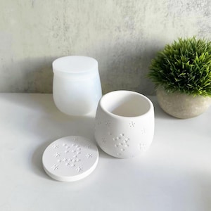 Puede incluir: Dos recipientes de cerámica blanca con un diseño floral. El recipiente más grande tiene una tapa. El recipiente más pequeño tiene una tapa que está separada.