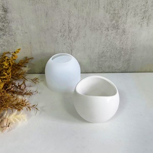 Könnte beinhalten: Zwei weiße Keramik-Blumentöpfe, einer rund und einer mit einer geschwungenen, asymmetrischen Form. Beide Töpfe sind glatt und haben eine matte Oberfläche.