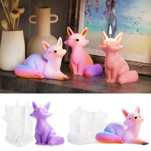 Peut inclure: Quatre bougies en forme de renard, deux roses, une violette et une avec une queue arc-en-ciel. Les bougies sont posées sur une surface en bois. Il y a aussi deux moules en silicone blanc pour faire des bougies en forme de renard.