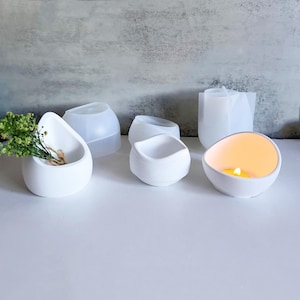 May include: Five white silicone molds for making concrete planters and candle holders. The molds are in various shapes, including a teardrop, a round bowl, and a rectangular bowl. One of the molds has a lit candle inside.