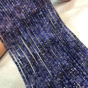 May include: Multiple strands of faceted, deep blue gemstone beads arranged in parallel lines. Each bead features a small, reflective surface that catches light, creating a shimmering effect across the collection of dark indigo-colored circular stones.