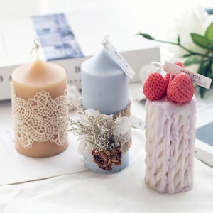 May include: Three decorative candles with different designs. The first candle is beige with a white lace design. The second candle is light blue with a dried flower arrangement. The third candle is a light purple with a textured design and two red strawberry decorations. Each candle has a small white label with the text "MAN YUE" printed on it.