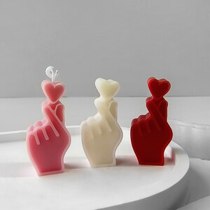 May include: Three small hand-shaped candles, each with a heart on the index finger. The candles are pink, white, and red.