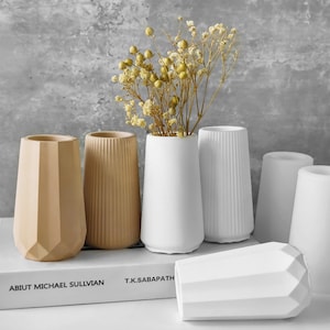 May include: Five different ceramic vases in various shapes and sizes. Two are beige, two are white, and one is a light gray. The vases are arranged on a white surface with a book titled "Abut Michael Sullivan" and "T.K. Sabapathi" visible in the background. Dried flowers are in one of the white vases.