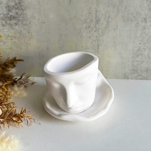 Puede incluir: Maceta de cerámica blanca con forma de cara con los ojos cerrados y una nariz. La maceta tiene un platillo.