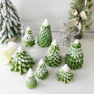 May include: A set of nine green and white Christmas tree candles. The candles are arranged on a white surface and are all different sizes. The candles are decorated with a white snowy top.