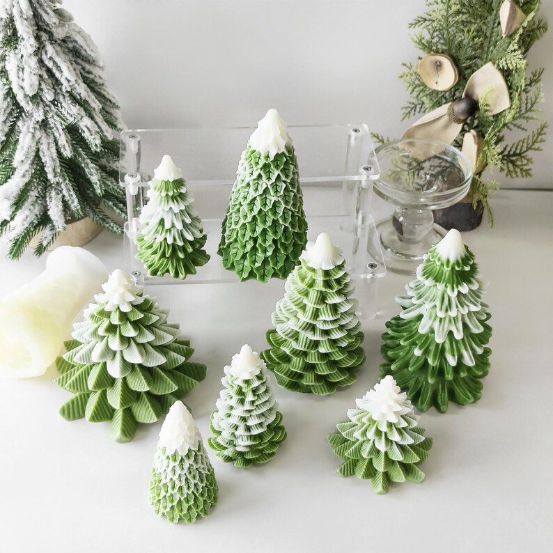 May include: A set of nine green and white Christmas tree candles. The candles are arranged on a white surface and are all different sizes. The candles are decorated with a white snowy top.