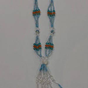 Op de afbeelding: Een lange kralenketting met lichtblauwe, doorzichtige, turquoise en oranje glaskralen in een meerstrangig patroon. De ketting eindigt in een kwastontwerp met vijf bungelende witte kaurischelpen bevestigd aan kralenstrengen.