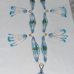 May include: A long beaded necklace featuring multiple strands of light blue and clear glass beads. The design includes sections of orange accent beads, decorative side tassels, and a central pendant finished with a blue bead and cowrie shells.