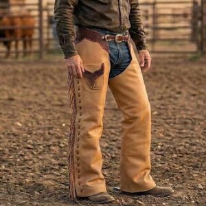 May include: Tan leather chaps with fringe detailing. The chaps have a brown leather yoke and a pocket with a decorative emblem. They are worn with a brown leather belt and blue jeans.