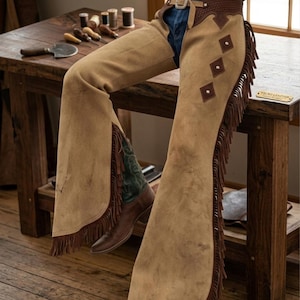May include: Tan leather chaps with brown fringe and diamond-shaped accents. The chaps are worn with a brown belt and blue jeans. Brown cowboy boots with green and brown detailing are visible. The chaps are resting on a wooden table.