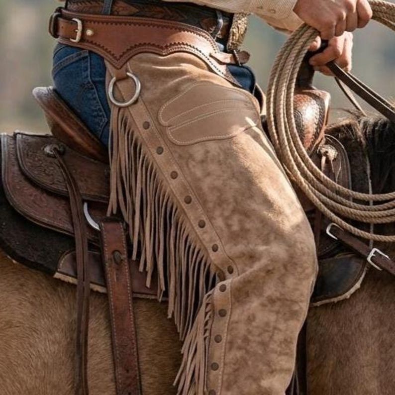 Close-up of a cowboy's upper hip and waist while riding. Features roughout suede leather chaps with side fringe, metal studs, and a decorative patch pocket. Includes a hand-tooled floral leather belt with buckles, worn over blue jeans, on a detailed western saddle.
