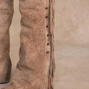 Close-up of the lower leg of roughout suede leather chaps. Features long leather side fringe and a row of dark metal studs along the seam. The bottom cuff rests over a distressed brown leather cowboy boot on a neutral studio background.