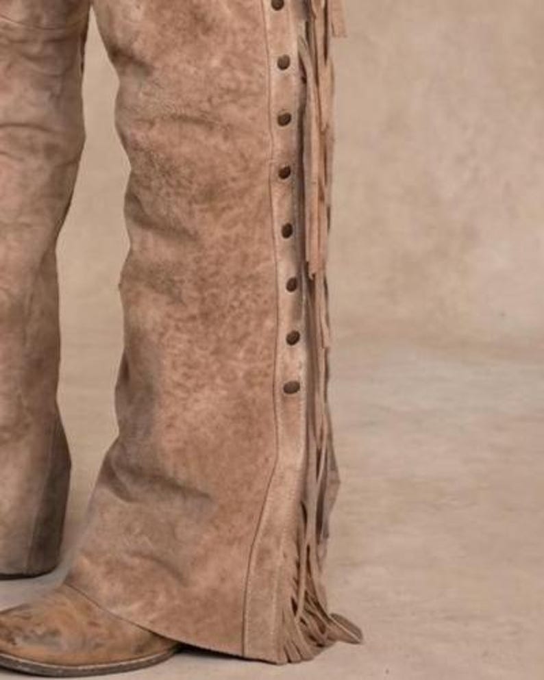 Close-up of the lower leg of roughout suede leather chaps. Features long leather side fringe and a row of dark metal studs along the seam. The bottom cuff rests over a distressed brown leather cowboy boot on a neutral studio background.