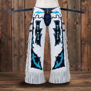 May include: White leather chaps featuring black and bright blue geometric overlays, silver-toned concho accents, and long white fringe along the outer legs. The chaps include adjustable black leather straps with metal buckles at the waist and thighs.