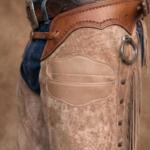 Close-up profile of roughout suede leather chaps worn over blue jeans. Features include a hand-tooled floral leather waistband, a pocket on the thigh, side fringe, a silver ring attachment, and a tooled belt with a silver buckle.