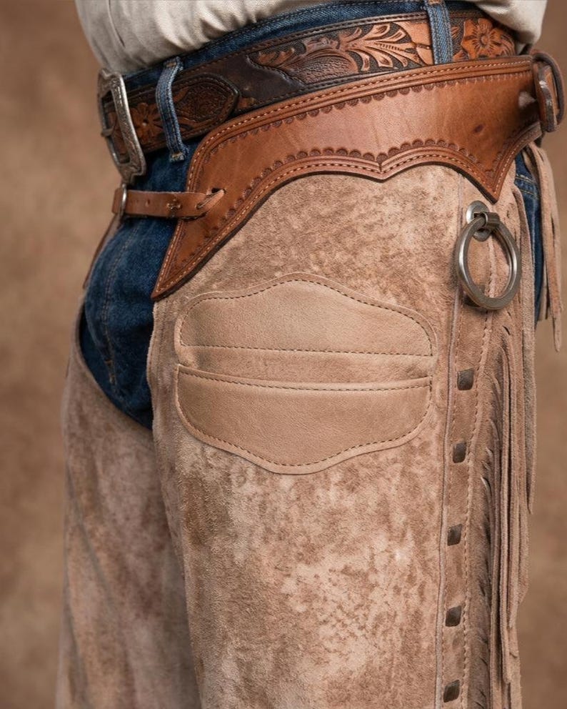 Close-up profile of roughout suede leather chaps worn over blue jeans. Features include a hand-tooled floral leather waistband, a pocket on the thigh, side fringe, a silver ring attachment, and a tooled belt with a silver buckle.