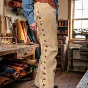 Peut inclure: Chaps en cuir beige avec franges et boutons argentés. Les chaps ont une ceinture en cuir marron. L'image est prise dans un atelier avec des rouleaux de cuir et des outils. Porté avec une chemise en jean.