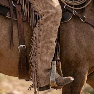 Close-up of a cowboy's leg in roughout suede leather chaps while riding a horse. The light brown chaps feature long side fringe and small dark studs along the seam. The foot rests in a metal stirrup on a tooled leather saddle.