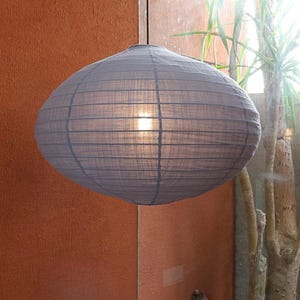 Puede incluir: Una gran lámpara de papel gris, de forma ovalada, cuelga de un cable negro. La lámpara está iluminada desde dentro, emitiendo un brillo suave. El fondo presenta una pared naranja y algo de vegetación.