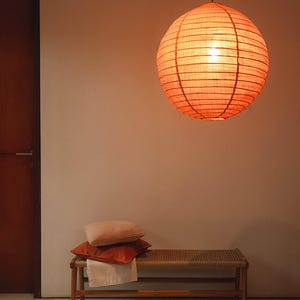 Puede incluir: Una linterna redonda de papel naranja cuelga del techo, iluminando un banco con cojines y un paño doblado. El banco tiene un asiento tejido y patas de madera. Dos cojines naranjas y un cojín rosa están apilados en el banco. Una puerta de madera está a la izquierda.