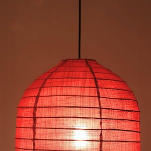 May include: A red, dome-shaped pendant light fixture. The shade is made of a textured, semi-transparent material, with horizontal and vertical dark lines creating a grid pattern. A black cord suspends the light from above, and the light source is visible inside.