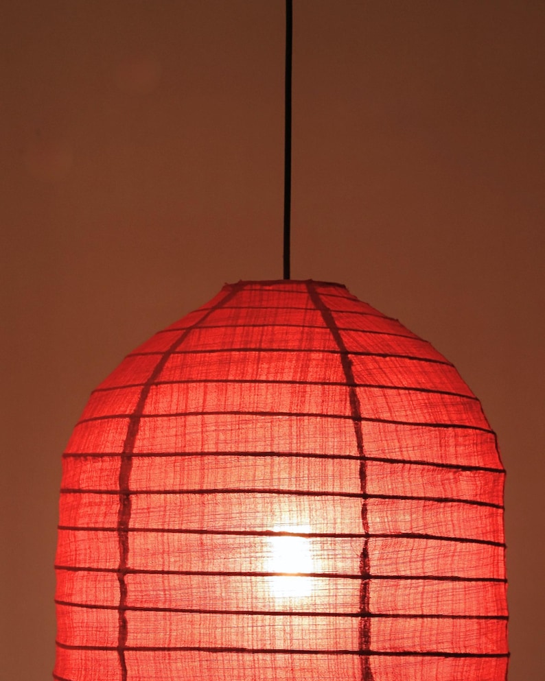 May include: A red, dome-shaped pendant light fixture. The shade is made of a textured, semi-transparent material, with horizontal and vertical dark lines creating a grid pattern. A black cord suspends the light from above, and the light source is visible inside.