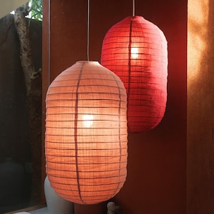 May include: Two hanging paper lanterns in shades of red and peach. The lanterns have a cylindrical shape with horizontal lines and are illuminated from within, creating a warm glow. The background is a dark red wall.