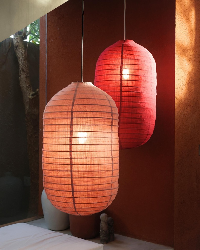 May include: Two hanging paper lanterns in shades of red and peach. The lanterns have a cylindrical shape with horizontal lines and are illuminated from within, creating a warm glow. The background is a dark red wall.