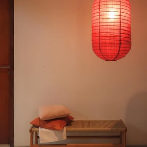 May include: A red paper lantern hangs from the ceiling, illuminating the space. Below, a wooden bench with a woven seat holds two orange pillows and a white cloth. The scene is bathed in warm light, creating a cozy atmosphere.
