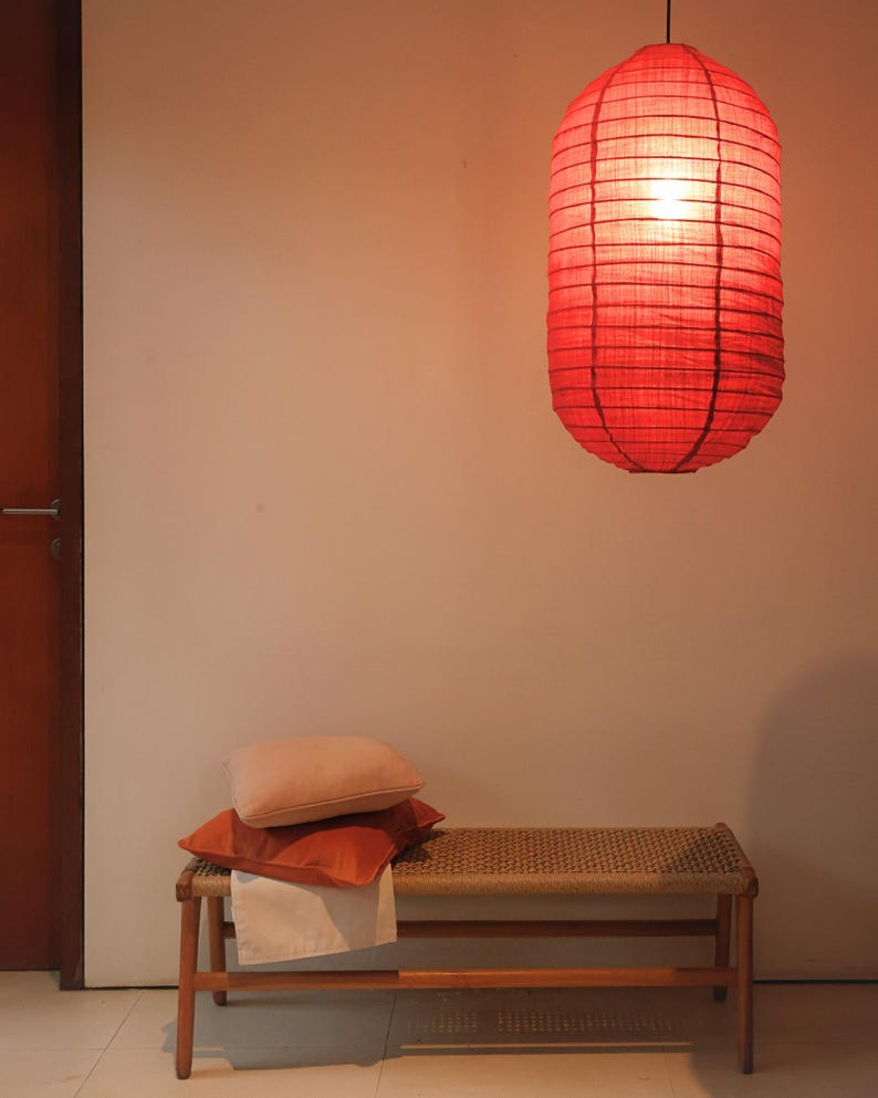 May include: A red paper lantern hangs from the ceiling, illuminating the space. Below, a wooden bench with a woven seat holds two orange pillows and a white cloth. The scene is bathed in warm light, creating a cozy atmosphere.