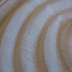 May include: Close-up of a cream-colored fabric with a subtle woven texture. The fabric is draped in soft, flowing folds, creating a sense of movement and depth. The word "TALANA" is printed in a light gray color on the right side of the fabric.