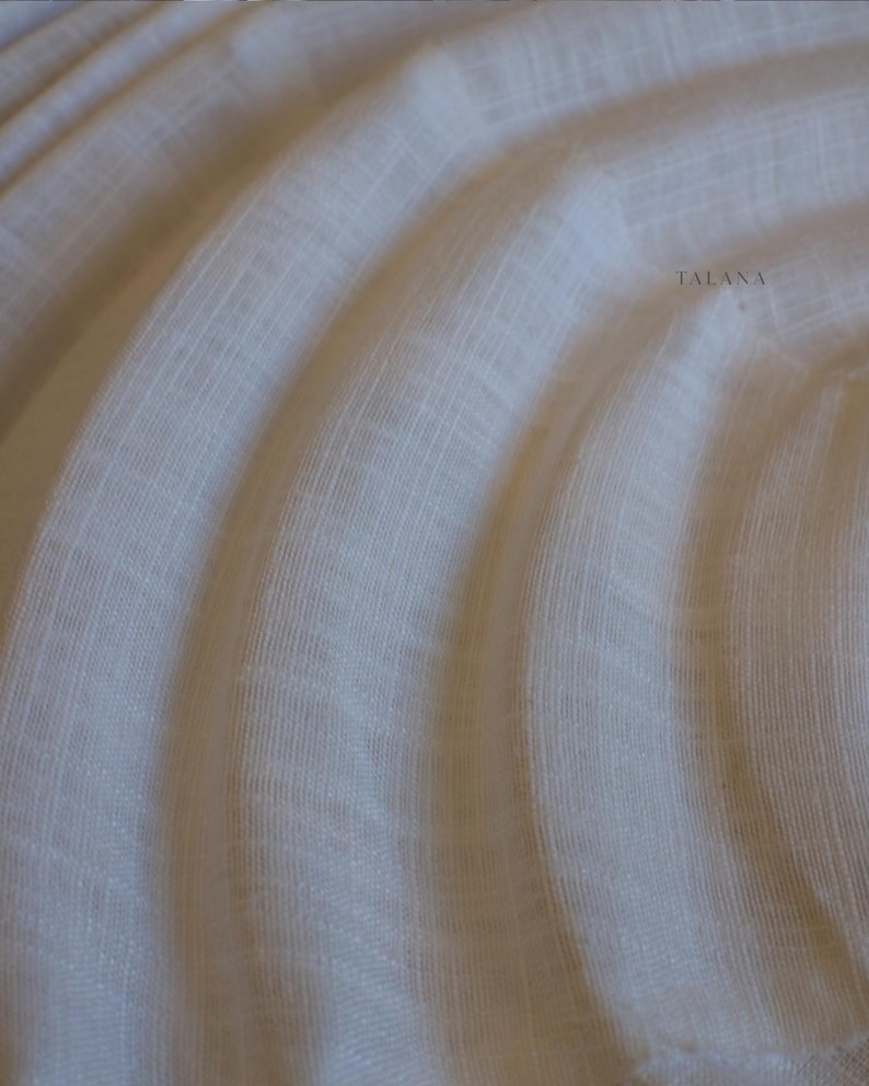 May include: Close-up of a cream-colored fabric with a subtle woven texture. The fabric is draped in soft, flowing folds, creating a sense of movement and depth. The word "TALANA" is printed in a light gray color on the right side of the fabric.